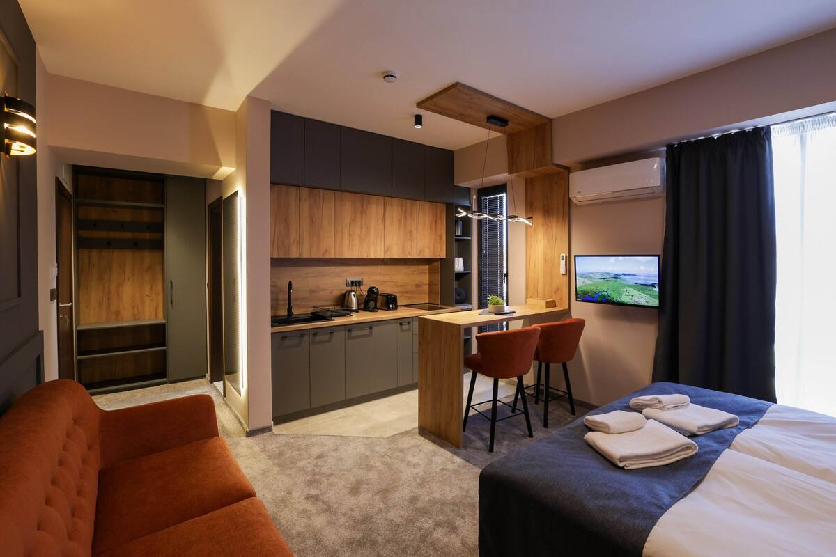A modern hotel room with a bed, a sofa, a small kitchen area, and a TV.