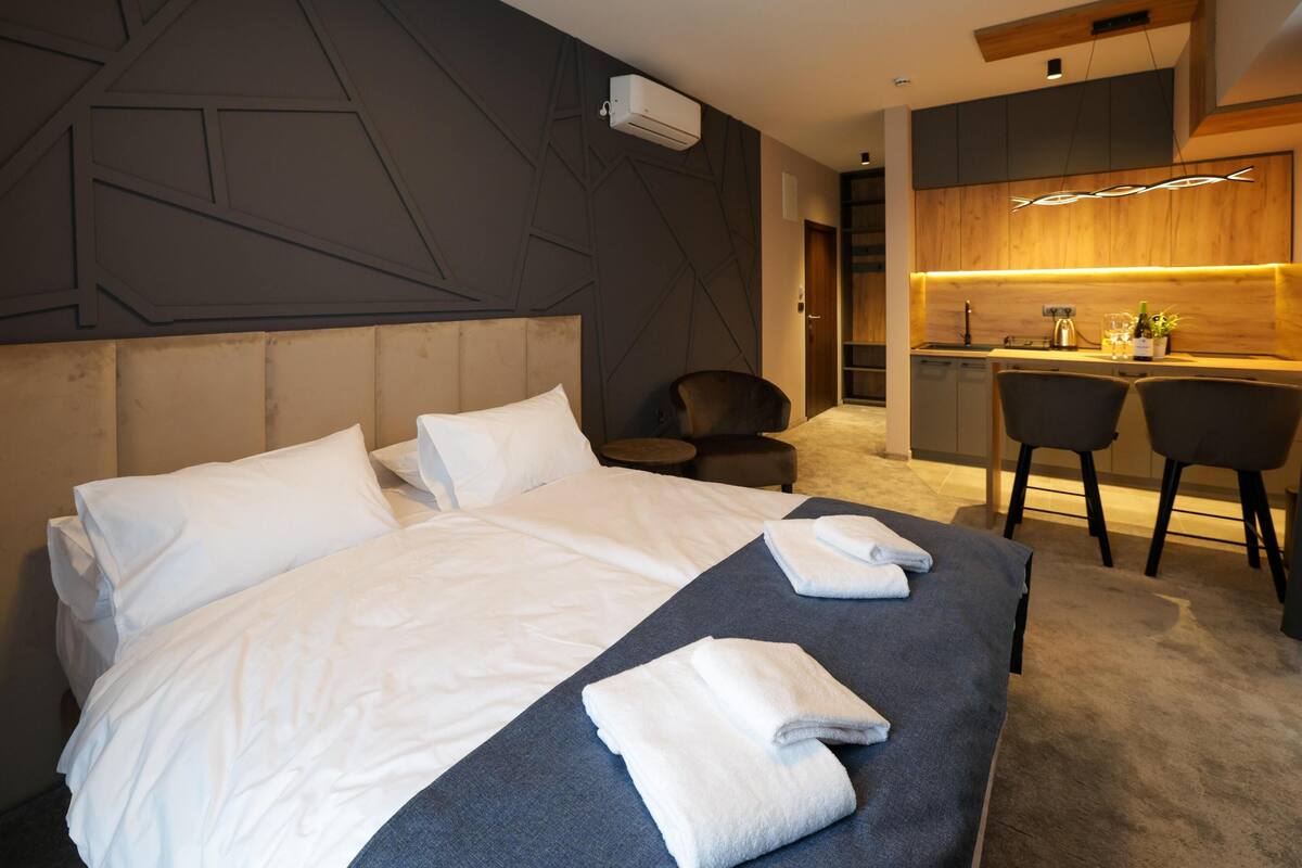 A modern hotel room with a large bed, a small desk, and a kitchenette.