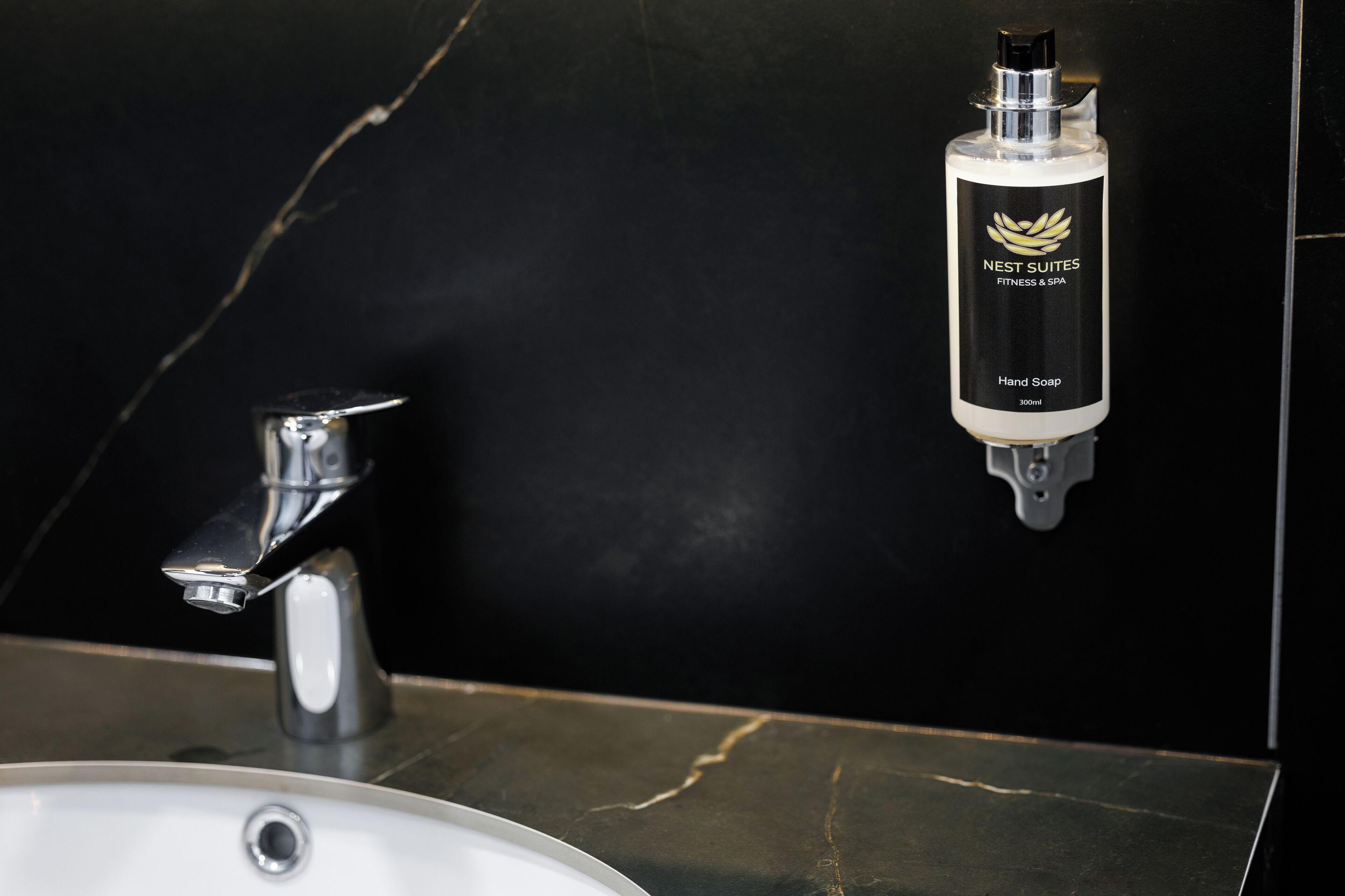 Un baño con pared de mármol negro, un lavamanos blanco y dispensador de jabón.