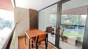 Un balcón con una mesa y sillas de madera, un ventanal de vidrio que da a un espacio al aire libre con un automóvil y vegetación.
