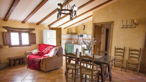 A cozy living room with a sofa, a dining table with chairs, a kitchen area with a refrigerator and microwave, and a window with curtains.