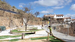 A playground with green artificial grass and sand areas, surrounded by stone walls and trees.