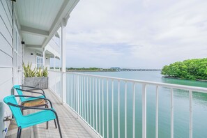 A balcony with two turquoise chairs, a concrete planter with green plants, and a view of a calm body of water with a pier in the distance.