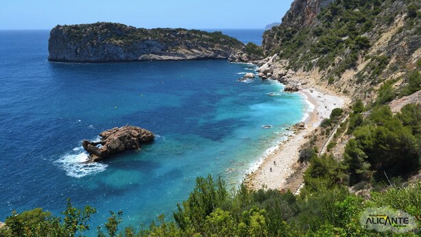 A coastal landscape with a rocky beach, clear blue sea, and a mountainous backdrop.