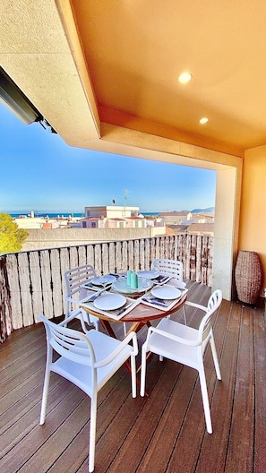 Una terraza de madera con una mesa redonda para dos, sillas blancas y vista a la ciudad y al mar de fondo.