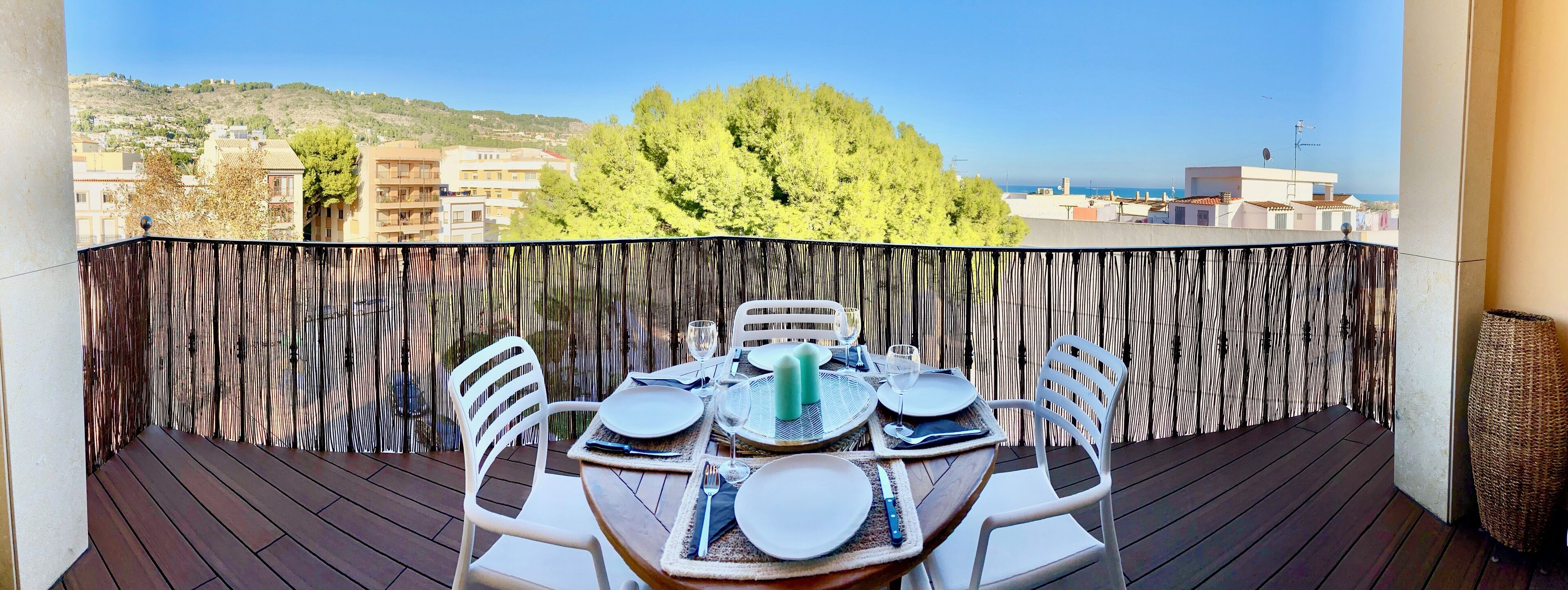 Ein Balkon mit Holzboden, weißen Stühlen und einem für das Essen gedeckten Tisch. Eine Bambus-Umzäunung trennt den Balkon von der Aussicht auf Gebäude und Bäume.