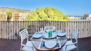 A balcony with a wooden floor, white chairs, and a table set for dining. A bamboo fence separates the balcony from the view of buildings and trees.