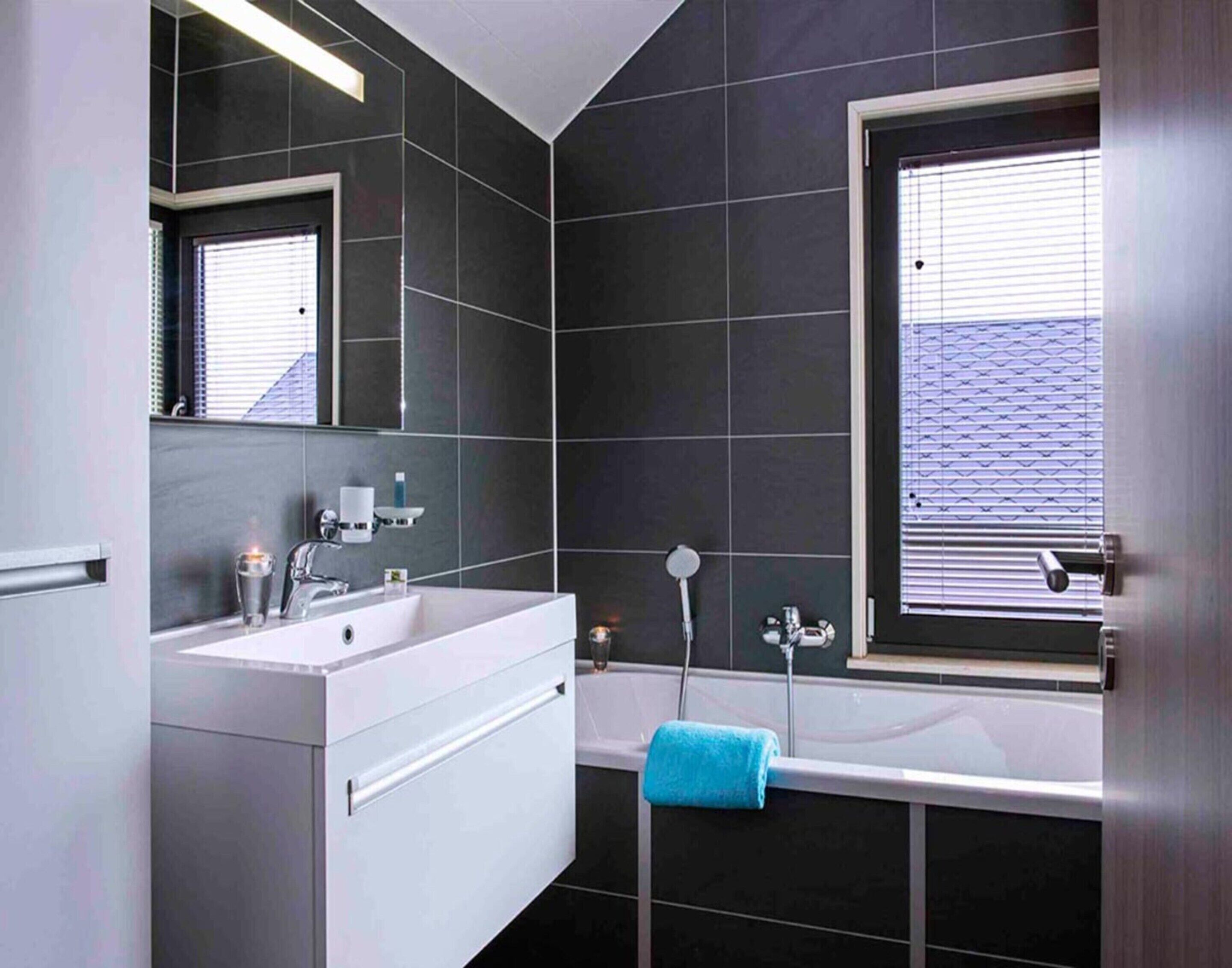 Une salle de bain moderne avec des carreaux foncés, un meuble-lavabo blanc, une baignoire et une fenêtre avec des stores.