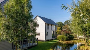 A modern house with a deck, surrounded by lush greenery and a pond. - Serene Luxe Lakeside Stay (Froidchapelle)