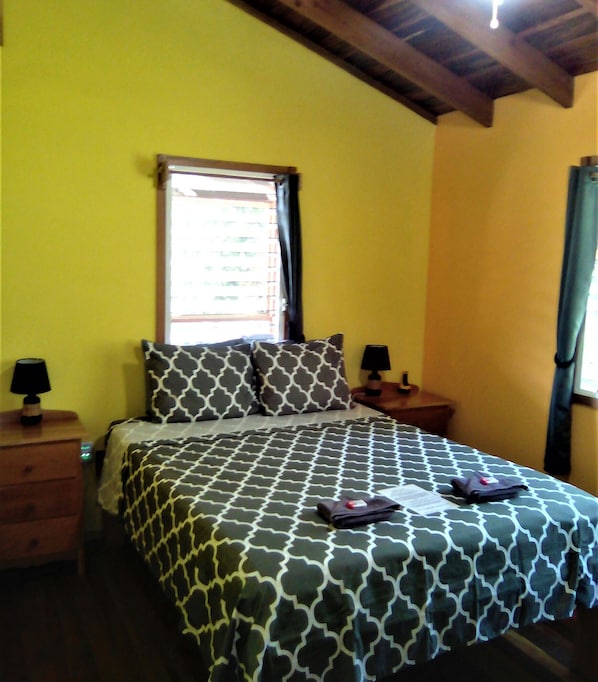 A bedroom with a bed, two nightstands, a window with blinds, and a ceiling with exposed beams.