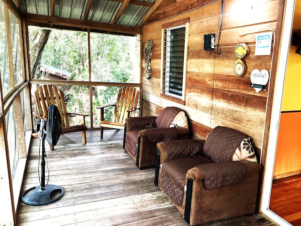 A cozy cabin interior with wooden walls, a wooden ceiling, a wooden floor, two wooden chairs, a brown sofa, a standing fan, and a window with a metal grill.