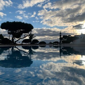 Un área de piscina tranquila con un reflejo nítido de árboles y un edificio lejano, envuelta en un ambiente sereno.