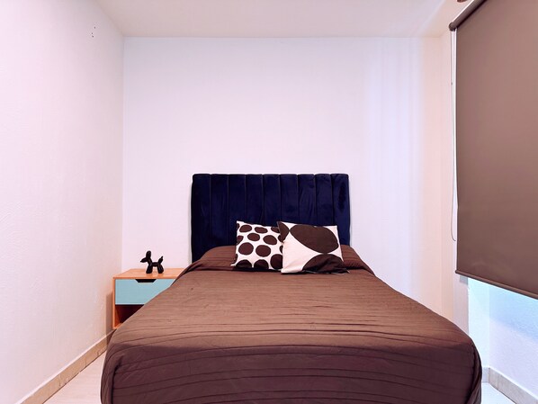 Une chambre d’hôtel étroite, avec un lit, une table de chevet et une fenêtre.
