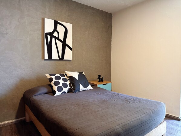 Un dormitorio con cama, almohadas, una mesita de noche y una obra de arte en la pared.