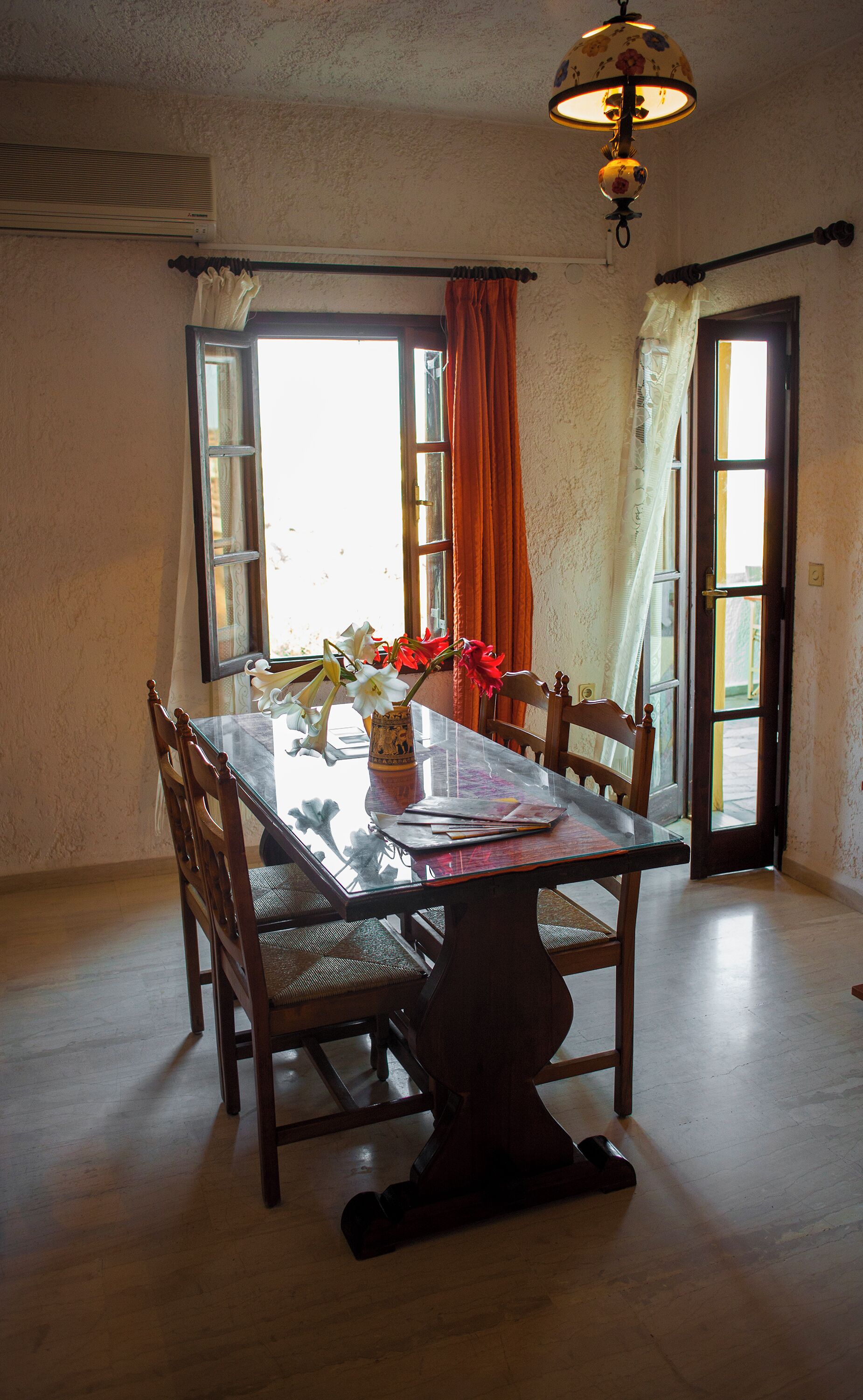 Een eetgedeelte met een glazen tafel, houten stoelen, een vaas met bloemen en een hanglamp.