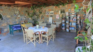 Une pièce rustique aux murs de pierre, avec un plafond en bois, une table ronde entourée de chaises, une bibliothèque et diverses plantes.