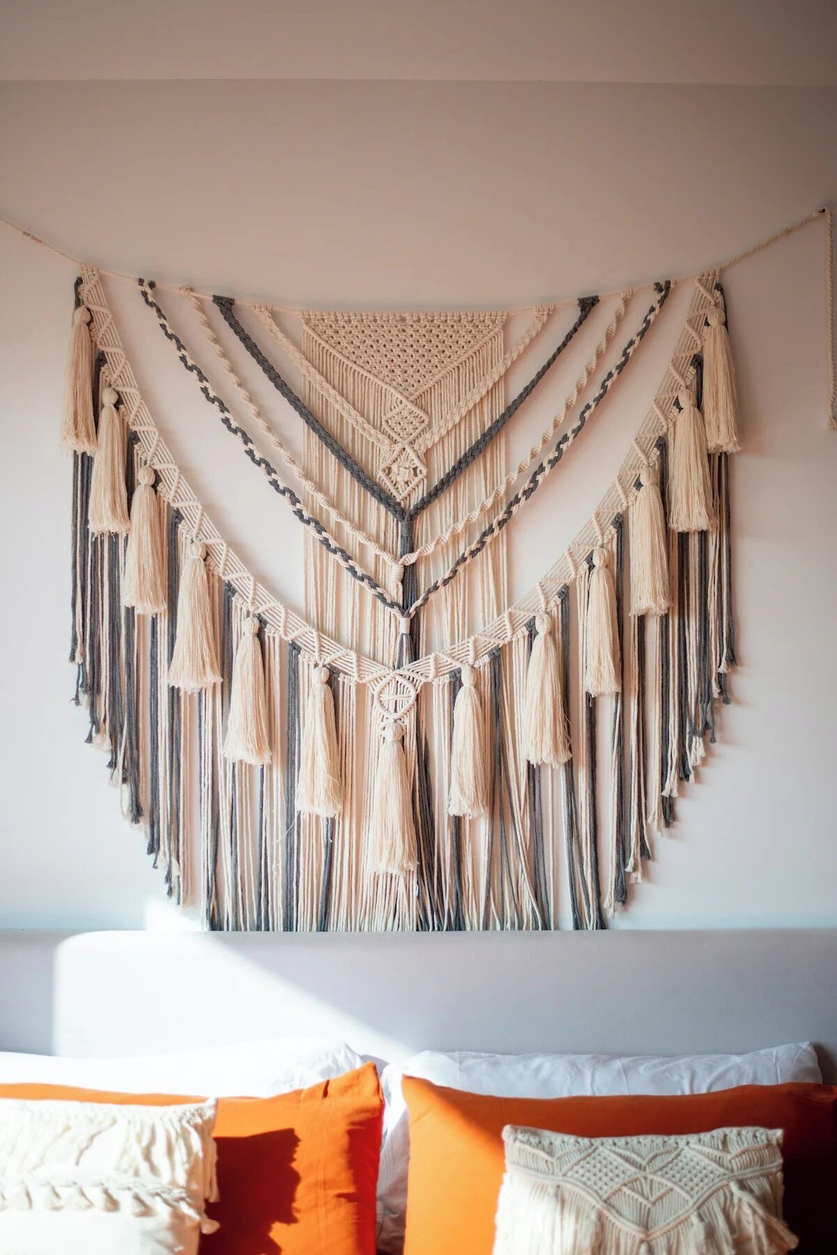 Un tapiz de macramé con flecos, una cama con almohadas blancas y naranjas, y una pared blanca lisa.