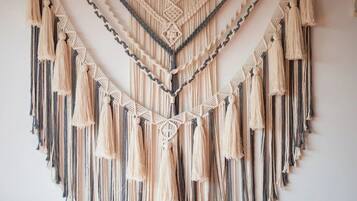 Un tapiz de macramé con flecos, una cama con almohadas blancas y naranjas, y una pared blanca lisa.