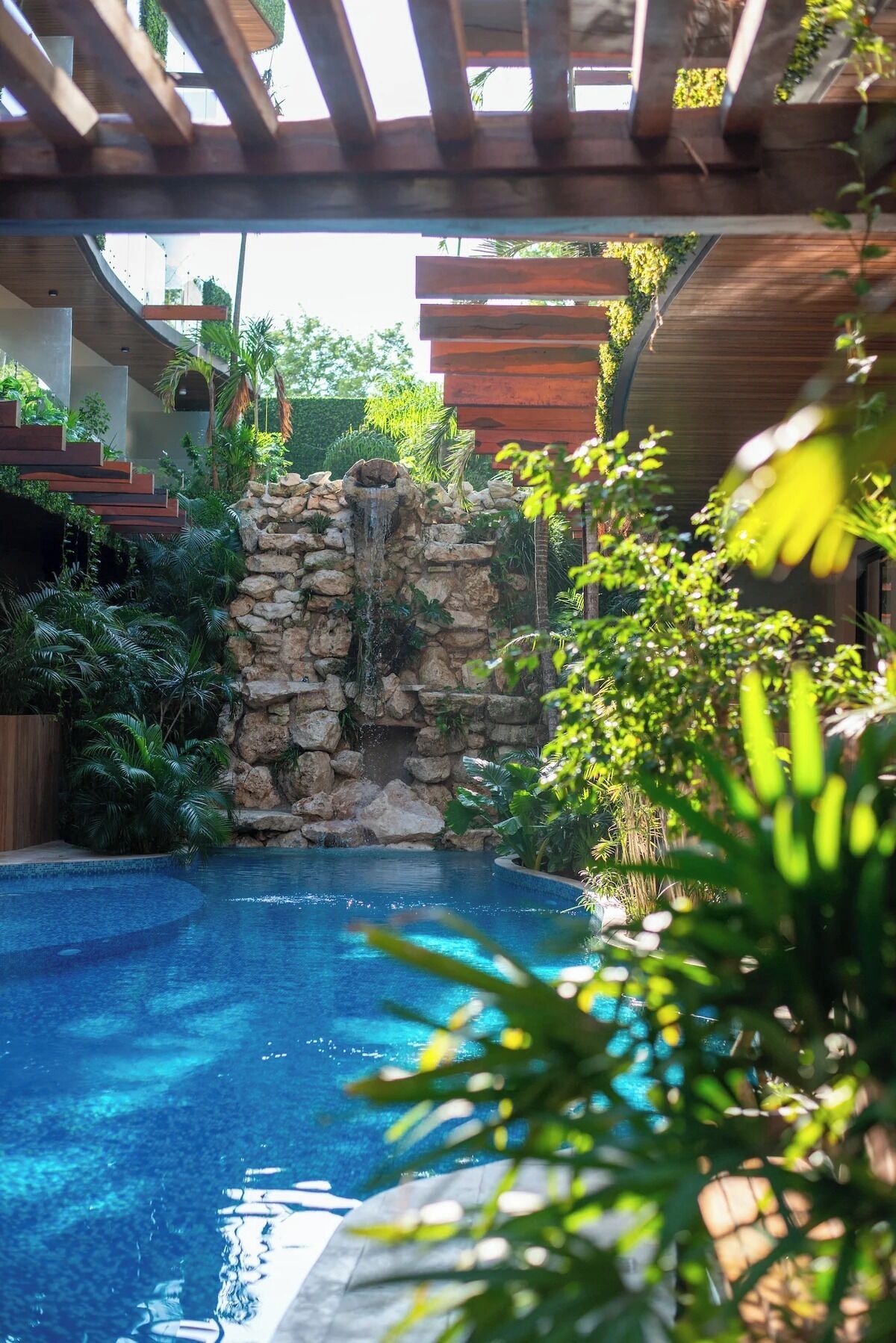 Una piscina con una cascada de piedra, rodeada de abundante follaje y una pérgola de madera.