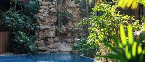 A pool with a stone waterfall feature, surrounded by lush greenery and a wooden pergola.