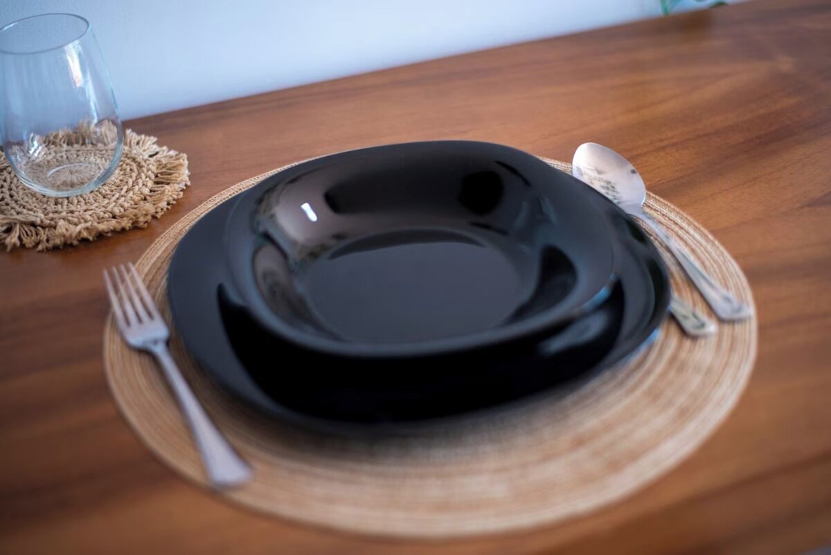 Una mesa de comedor con un plato negro, un tenedor, una cuchara y un vaso sobre un mantel tejido.
