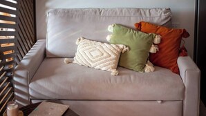 A sofa with three pillows, a small table with a candle and a notebook, and a window with blinds.