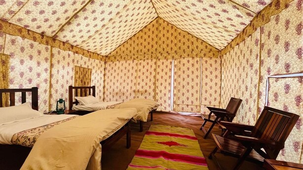 A room with two beds, a wooden chair, a small table, and a patterned wall. - Kumbh Travel (Prayagraj)