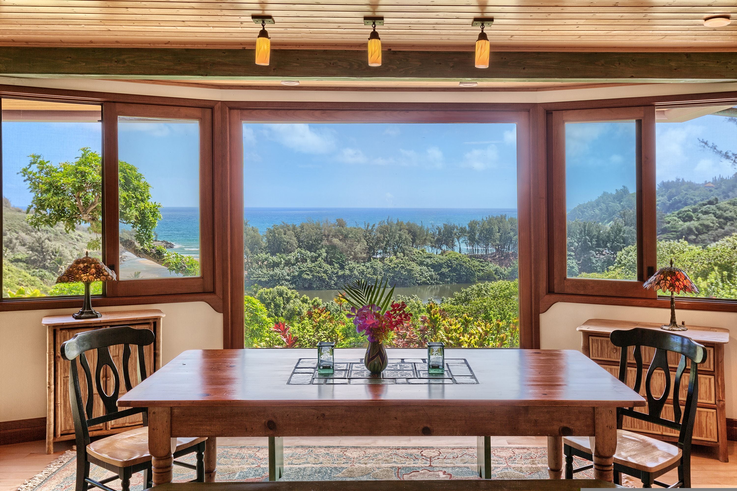 A dining area with a wooden table and chairs, large windows with a view of the ocean and greenery, and two lamps on the windowsill.