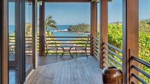 A wooden deck with a table and chairs, overlooking a coastal view with palm trees and a clear sky. - Ikena Lani in Kilauea (Kilauea)