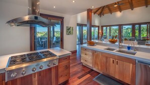 A modern kitchen with a stainless steel stove, wooden cabinets, and large windows offering a view of the outdoors. - Ikena Lani in Kilauea (Kilauea)