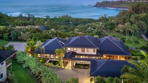 A large, multi-story house with a dark roof, surrounded by lush greenery and a view of the ocean. - Ikena Lani in Kilauea (Kilauea)
