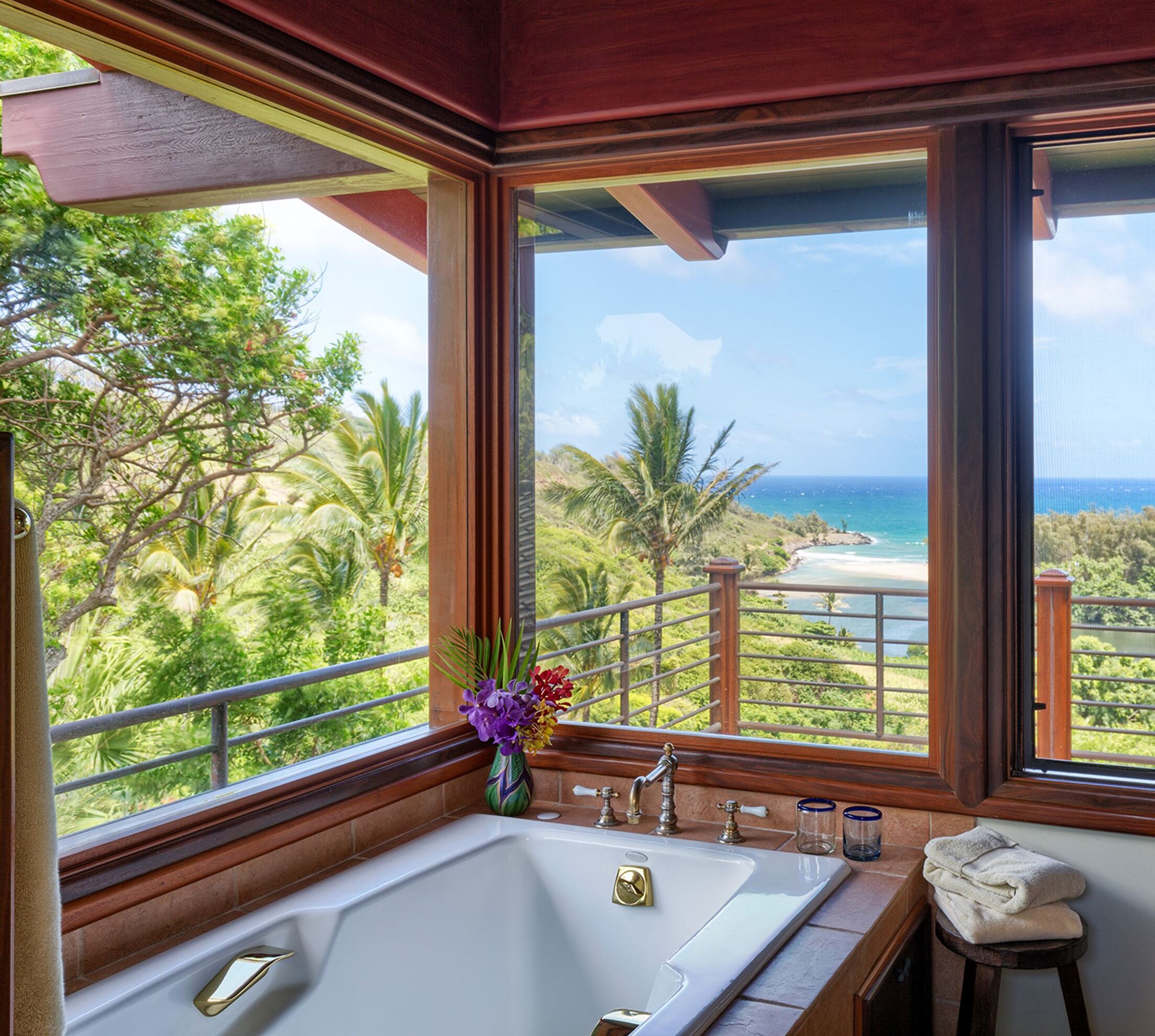 Un baño con una bañera grande, una ventana con vista a una playa y palmeras, y un florero con flores en el alféizar.