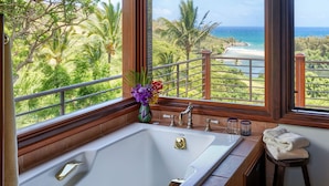 A bathroom with a large bathtub, a window with a view of a beach and palm trees, and a vase with flowers on the windowsill.