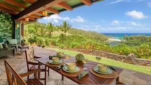 An outdoor dining area with a table set for four, overlooking a scenic view of a coastal landscape with hills and a beach.