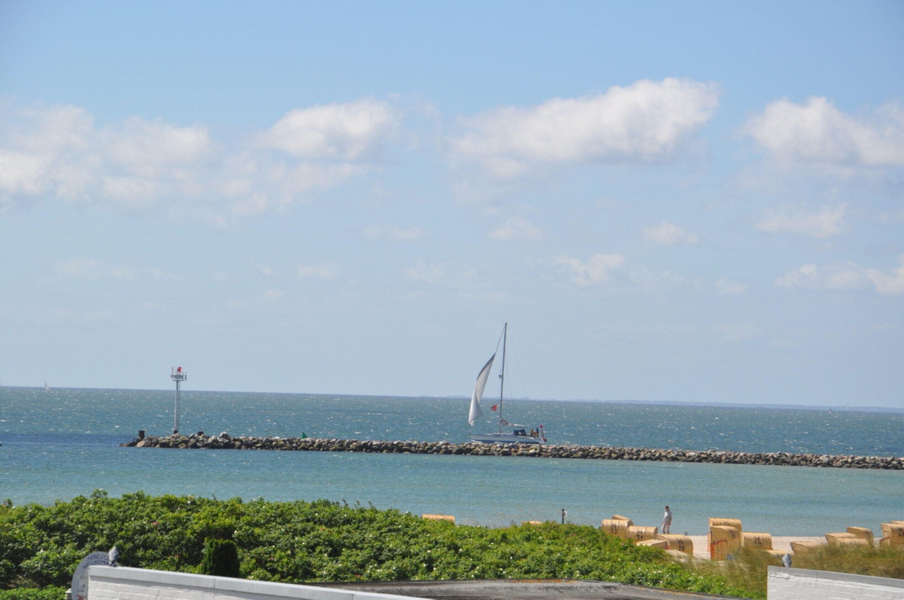A coastal view with a sailboat, a rocky breakwater, and a clear sky.