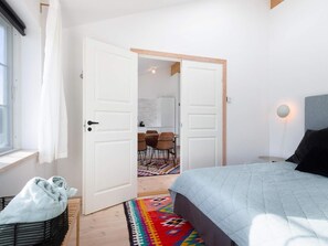 A bedroom with a bed, a bedside table, a lamp, a window with curtains, and a door leading to a dining area.