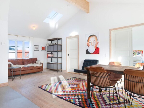 A spacious living area with a high ceiling, a wooden dining table with chairs, a colorful rug, a leather sofa, a bookshelf, and a large window with a view of buildings.
