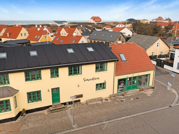 A building with a yellow facade and green windows, named 'Smedjen,' with a paved street in front and a view of the sea in the background.