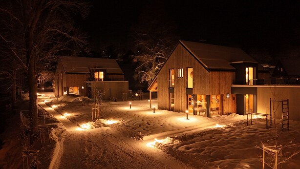 Paisaje nevado con edificios de madera, ventanas iluminadas y un sendero despejado.