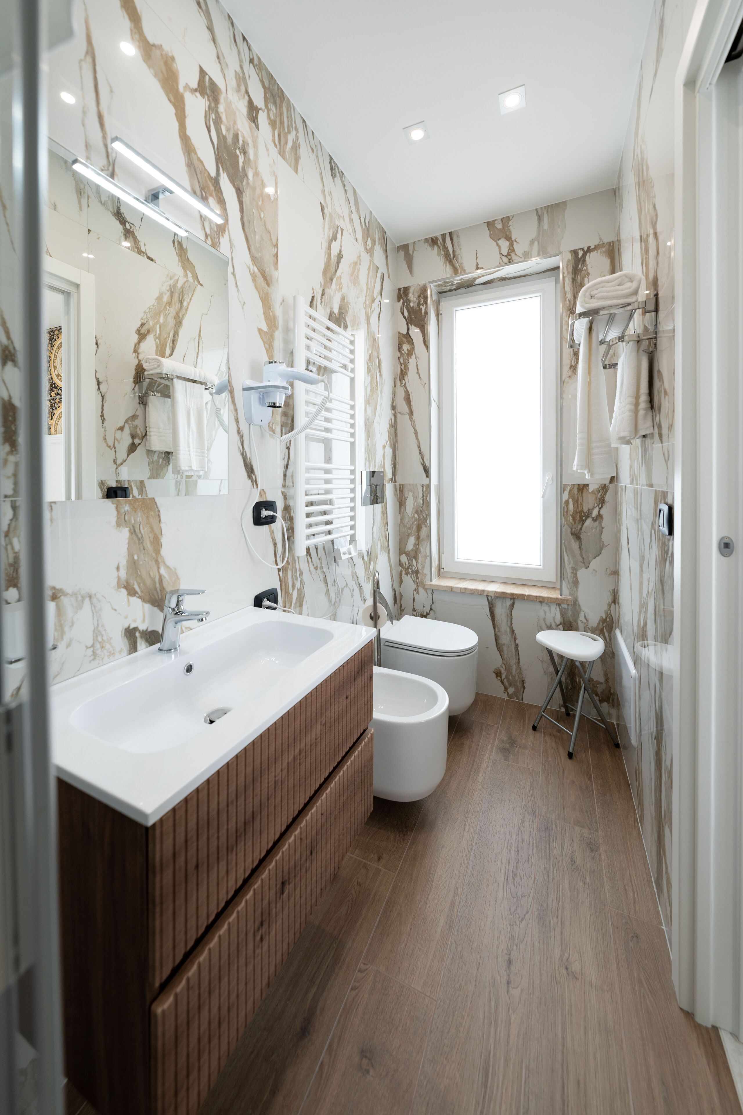 Un baño moderno con paredes de mármol, un mueble de tocador de madera, un lavabo grande, un inodoro y un ventanal que proporciona luz natural.