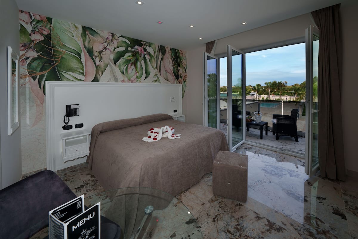 Hotelkamer met een bed, een glazen tafel, een menuhouder, een nachtkastje en uitzicht op een zwembad en terras.