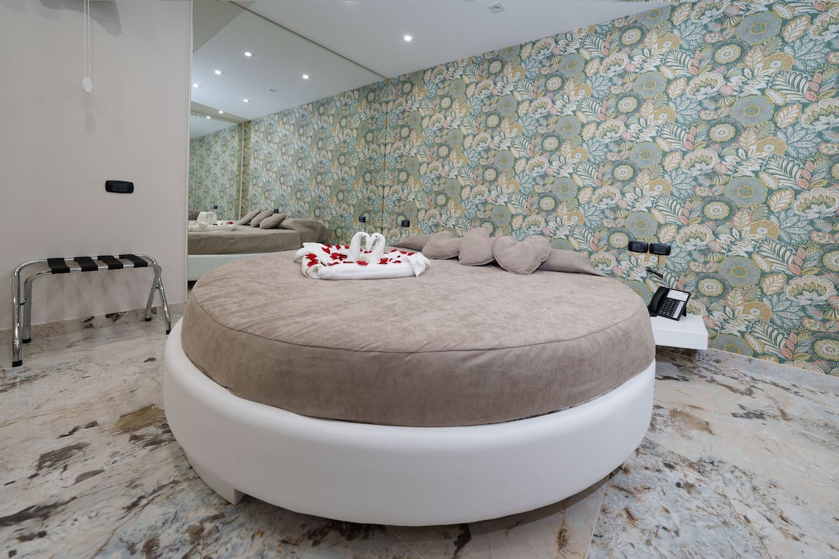 Een rond bed met een witte basis en beige beddengoed, een klein nachtkastje met een telefoon, en een wand met een bloemenpatroon.