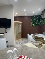 Una habitación de hotel moderna con televisor de pantalla plana, sauna de madera, bañera de hidromasaje blanca y zona de comedor con suelo de mármol.