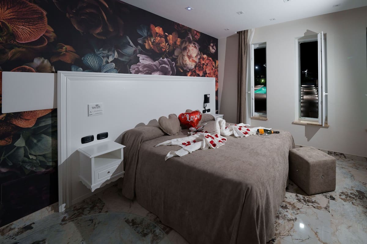 Een slaapkamer met een bloemenmural, een bed met een beige dekbedovertrek, een klein wit nachtkastje en een raam met gordijnen.