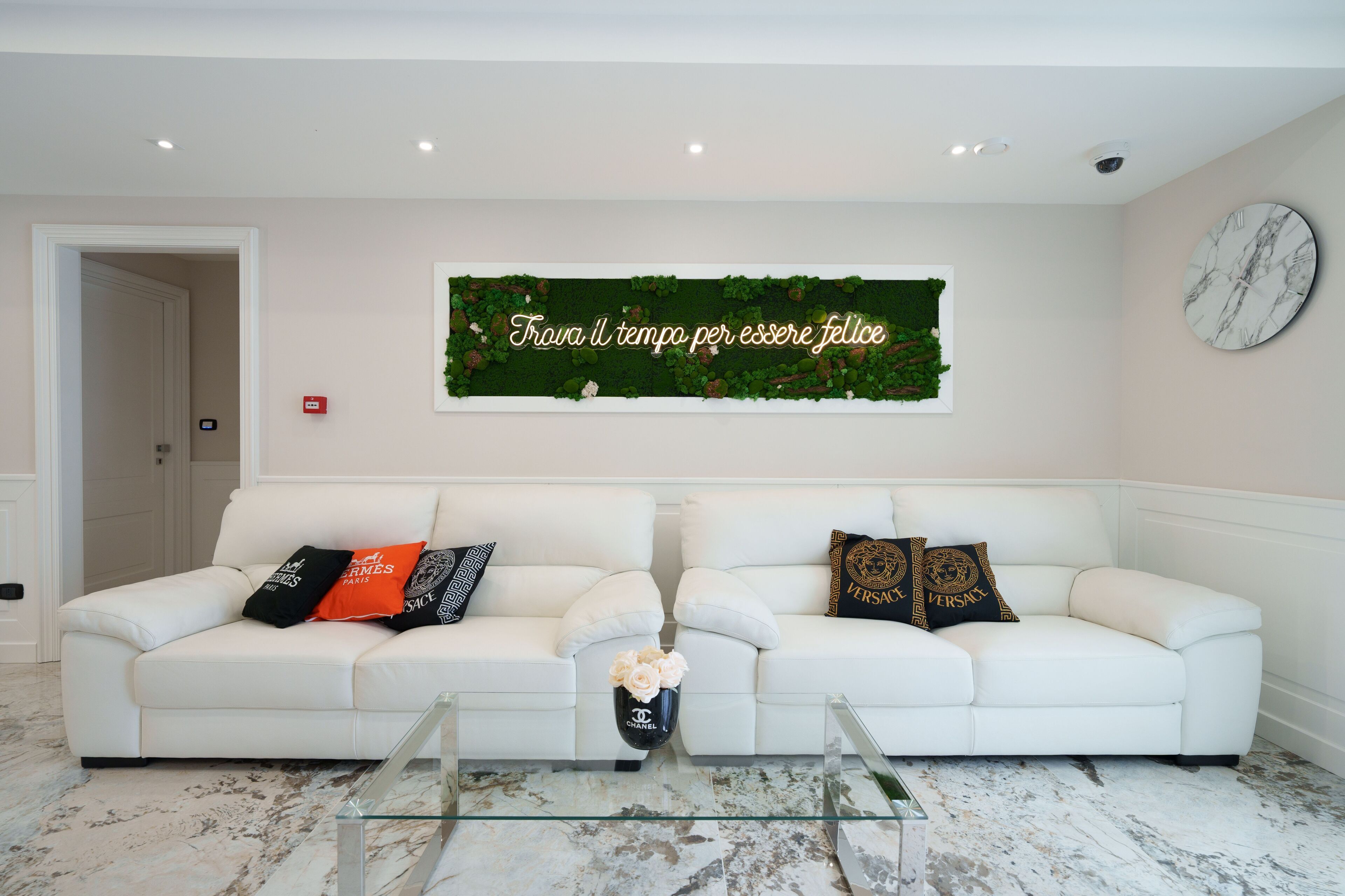 Ein modernes Wohnzimmer mit einem weißen Ledersofa, einem Glastisch und einer Wanddekoration mit einer Zimmerpflanze und einem motivierenden Zitat.