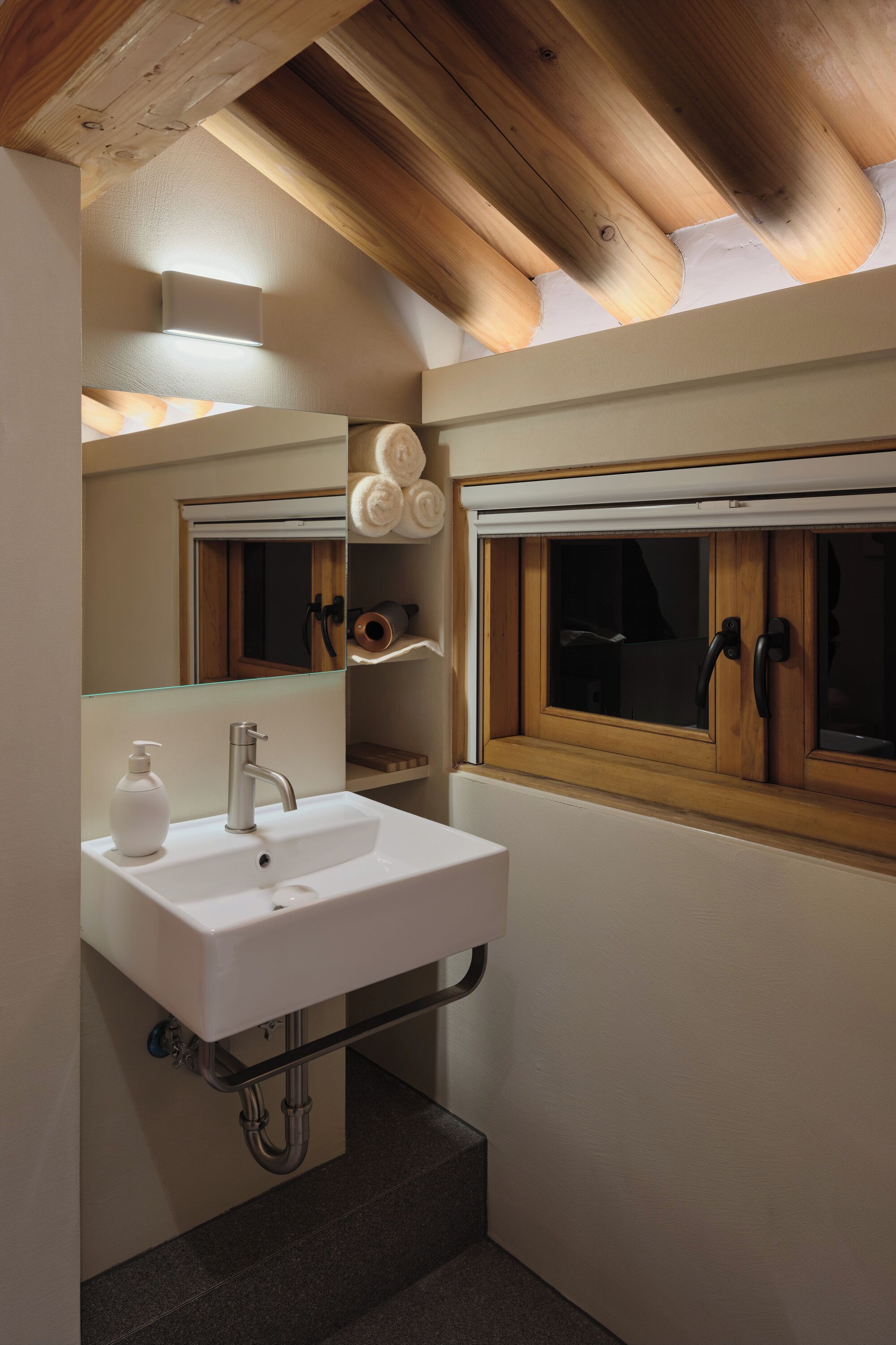 A bathroom with a white sink, a mirror, wooden beams on the ceiling, and a window with wooden shutters.