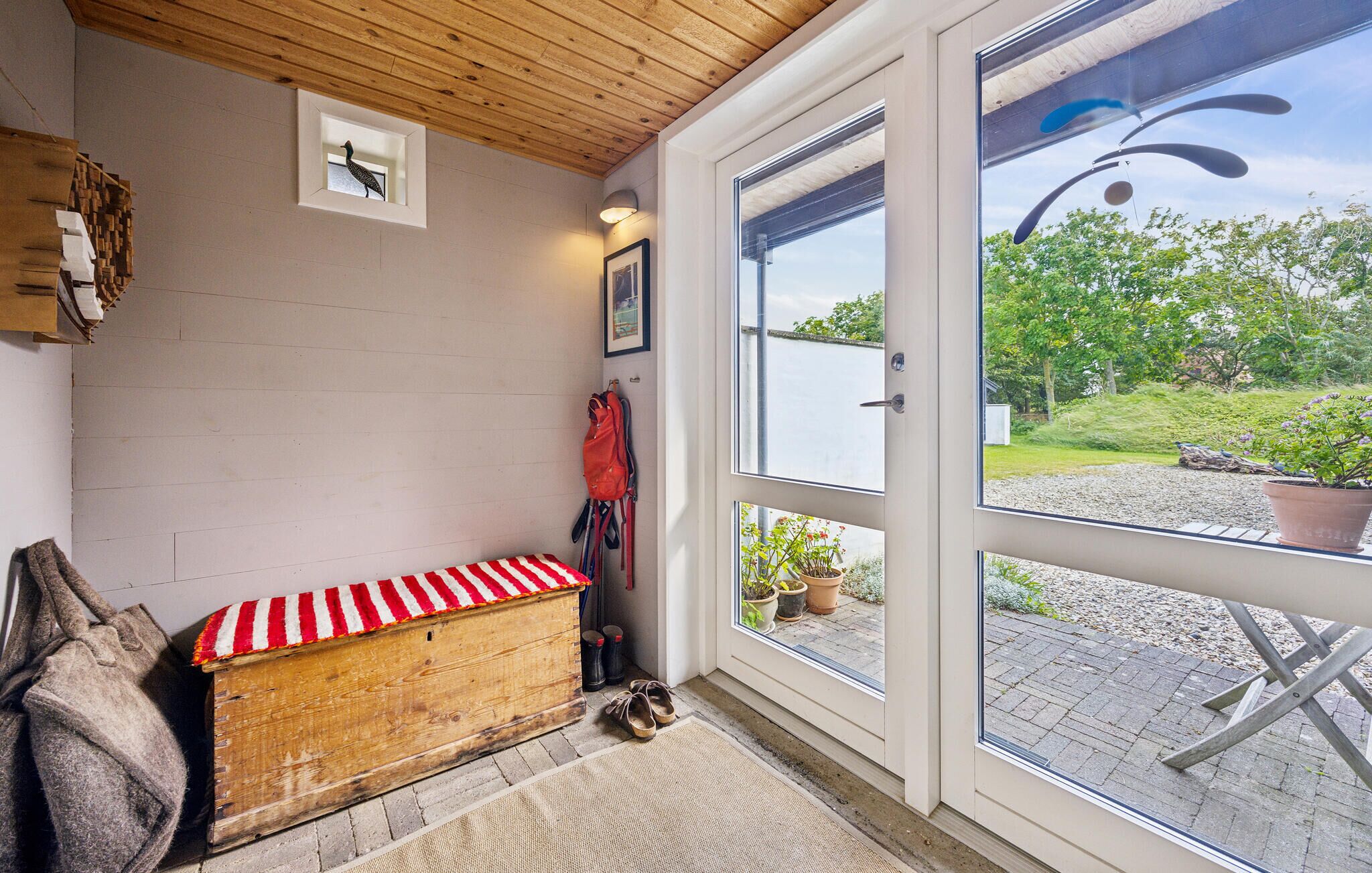 Une pièce chaleureuse avec un banc en bois, une petite fenêtre décorée d’un motif d’oiseau et une vue sur un jardin, visible à travers une porte vitrée.