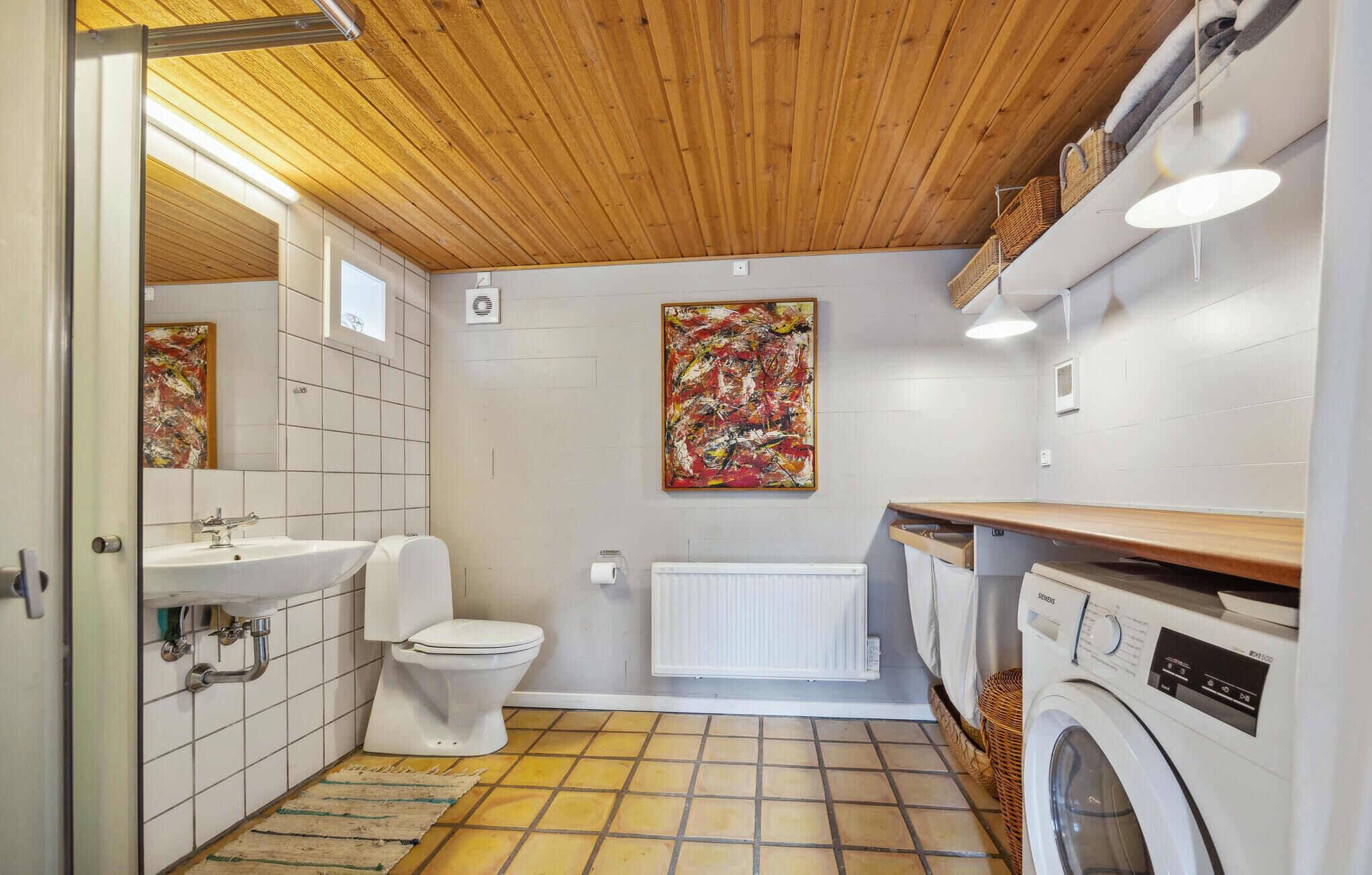 Een kleine badkamer met een houten plafond, een witte wastafel, toilet, radiator en wasmachine.