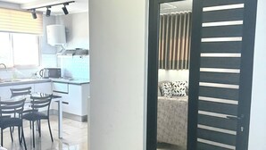 A modern living room with a grey sofa, a dining area with a table and chairs, and a kitchen with a water dispenser and a kettle.