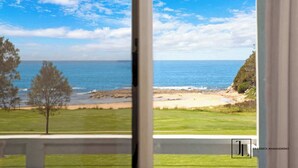 Blick durch ein Fenster auf einen Strand mit einer Rasenfläche und einem Baum, mit dem Logo von 'JL Property Management' unten rechts.